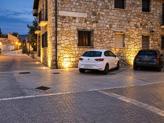 Fachada de piedra iluminada desde el pavimento, junto a la zona de aparcamiento.