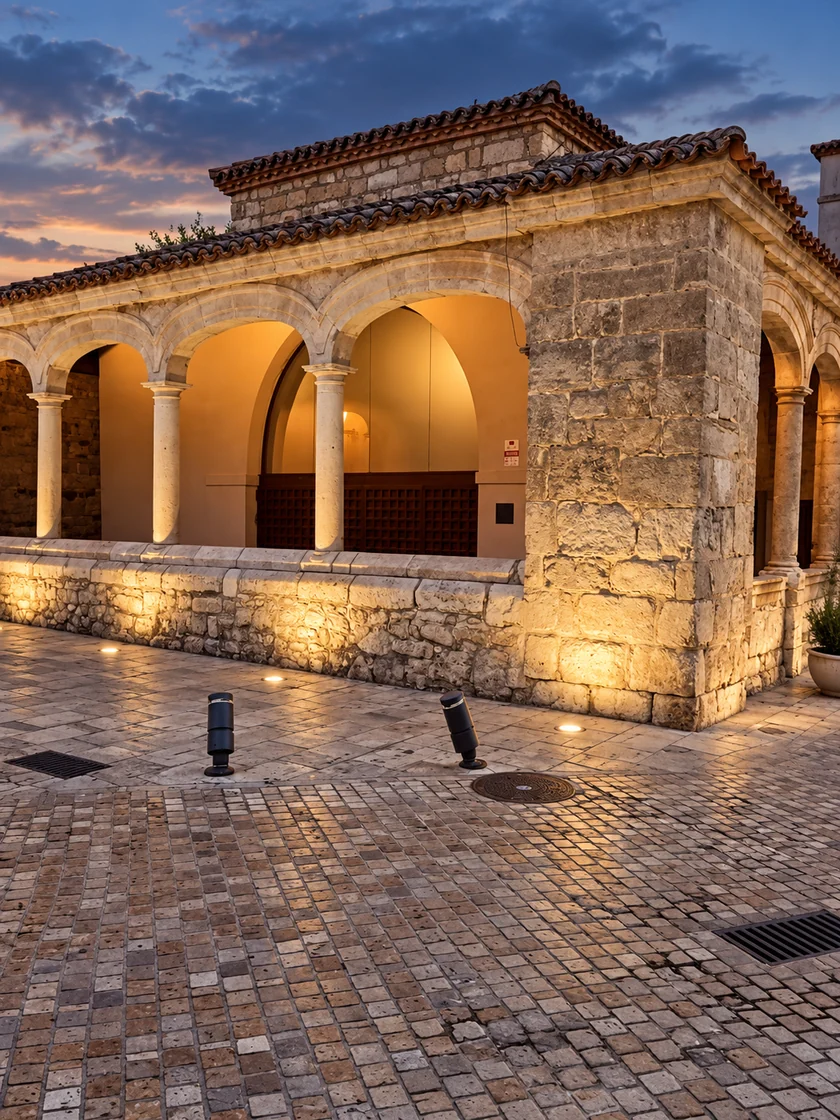 Vista en esquina del pórtico, con arcos y columnas de piedra iluminados al anochecer.