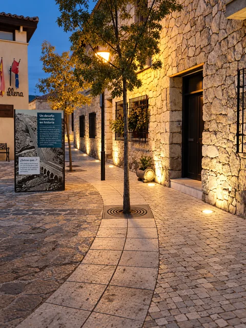 Puntos de luz a lo largo de una fachada de piedra, junto a puertas, ventanas y arbolado.
